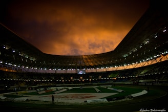 A large indoor stadium with an intricately designed roof is illuminated under a dramatic orange and black sky. The interior lighting creates a contrast with the natural lighting outside. A central track is visible with banners and advertisements surrounding it.
