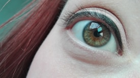 A close-up view of a human eye with brown and green iris colors. The surrounding eyelid is adorned with subtle makeup, and there is a reflection in the eye, likely of a window or some other structure. The person's hair, partially visible, appears reddish-brown.