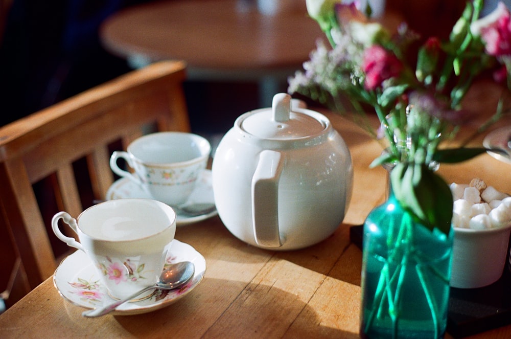 ピンクの花のセンターピースのクローズアップ写真の近くに茶色の木製のテーブルの上に白いセラミックのティーポットと2つの白いティーカップ