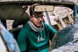A person with glasses and a green hoodie is sitting inside a damaged car. The interior is partially visible with the rearview mirror and some broken elements of the car. The background is blurred but consists of wooden debris.