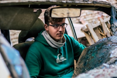 A person with glasses and a green hoodie is sitting inside a damaged car. The interior is partially visible with the rearview mirror and some broken elements of the car. The background is blurred but consists of wooden debris.
