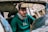 A person with glasses and a green hoodie is sitting inside a damaged car. The interior is partially visible with the rearview mirror and some broken elements of the car. The background is blurred but consists of wooden debris.