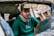 A person with glasses and a green hoodie is sitting inside a damaged car. The interior is partially visible with the rearview mirror and some broken elements of the car. The background is blurred but consists of wooden debris.