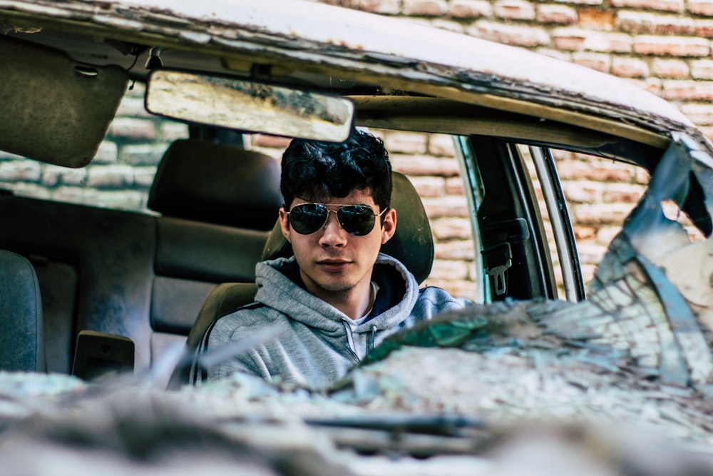 Hombre con sudadera con capucha gris y gafas de sol dentro de un coche destrozado