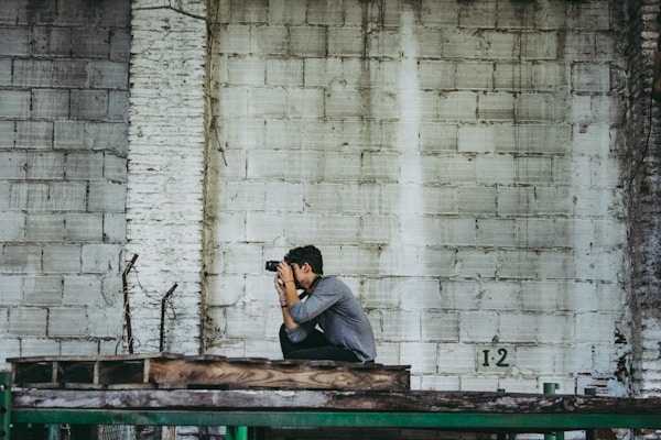 A person crouches on a wooden platform, holding a camera up to their face, poised to take a photo. They are positioned against a textured wall made of stacked concrete blocks with varying shades of grey. The setting appears industrial, as indicated by the rough materials and the wooden structure.