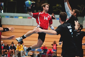 A handball player wearing a red jersey with the number 14 is airborne, dynamically reaching to throw a ball. His teammates and opponents in black jerseys are surrounding him. The scene is set in an indoor stadium with blurred spectators in the background.