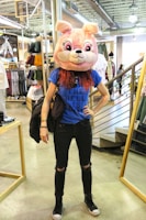 A person wearing a large, cartoonish animal head mask resembling a pink bear inside a clothing store. They are dressed in a blue T-shirt with writing on it, black ripped jeans, and black sneakers. The setting features clothing racks and a staircase.