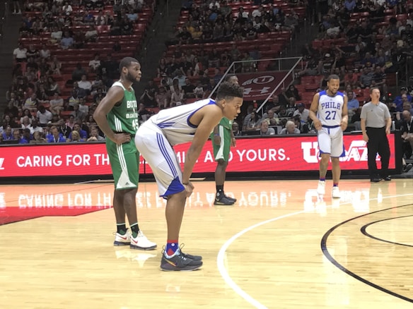 Two basketball teams are on the court during a game. One player in a green uniform from the Boston team is standing with hands on hips, while a player in a white and blue uniform from the Philadelphia team is bent forward slightly, appearing ready to make a move. Another player from the Philadelphia team, wearing number 27, is walking in the background. The court is surrounded by a crowded stadium with spectators, and a digital display screen shows advertisements and messages.