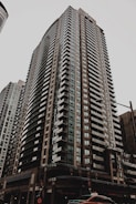 A tall residential skyscraper with modern architectural design, featuring numerous rows of windows and balconies. The building is situated in an urban environment with a cloudy sky overhead and a street scene at the base including a taxi cab and traffic lights.