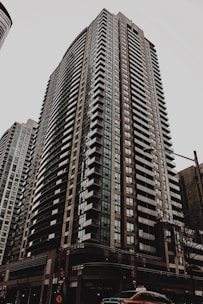 A tall residential skyscraper with modern architectural design, featuring numerous rows of windows and balconies. The building is situated in an urban environment with a cloudy sky overhead and a street scene at the base including a taxi cab and traffic lights.
