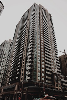 A tall residential skyscraper with modern architectural design, featuring numerous rows of windows and balconies. The building is situated in an urban environment with a cloudy sky overhead and a street scene at the base including a taxi cab and traffic lights.