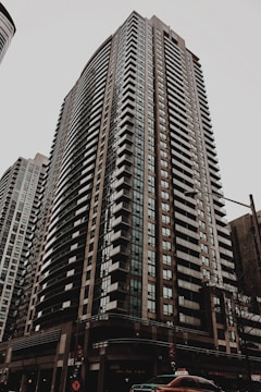 A tall residential skyscraper with modern architectural design, featuring numerous rows of windows and balconies. The building is situated in an urban environment with a cloudy sky overhead and a street scene at the base including a taxi cab and traffic lights.