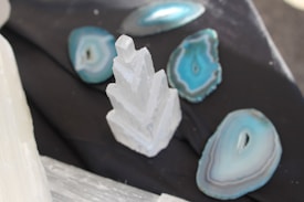 A white, geometric crystal structure is surrounded by slices of blue and teal agate geodes. The objects are placed on a dark, textured surface, creating a contrast between the vibrant geological specimens and the background.