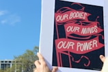 A person is holding a poster with red banners displaying the words 'OUR BODIES, OUR MINDS, OUR POWER' against a dark background. The sky is clear and blue, with the tops of some trees and a building visible in the background.