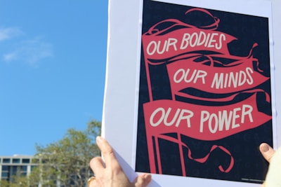 A person is holding a poster with red banners displaying the words 'OUR BODIES, OUR MINDS, OUR POWER' against a dark background. The sky is clear and blue, with the tops of some trees and a building visible in the background.