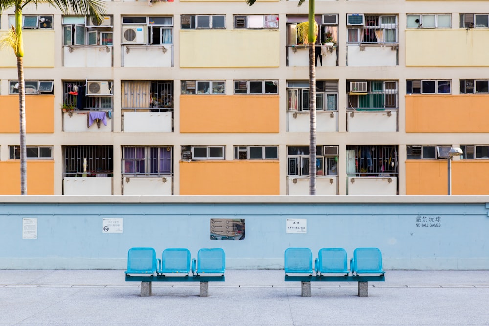 Dos sillas de pandilla azules de 3 asientos cerca de una pared de concreto azul y un edificio marrón