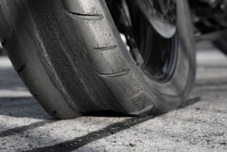 A close-up view of a worn motorcycle tire on a textured asphalt surface. The rubber appears smooth and the tread is nearly flattened, indicating significant use.