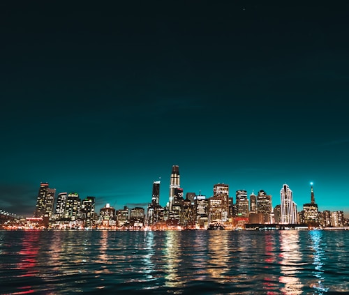 A nighttime cityscape of tall buildings with their lights illuminated against a dark blue to turquoise gradient sky. The city skyline reflects off the water in the foreground, creating a shimmering effect.
