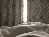 A softly lit bedroom with patterned curtains, featuring an intricate design of overlapping ovals and floral elements. The light from the window is partially obscured by the curtains, casting a gentle glow onto the rumpled white bedding in the foreground.