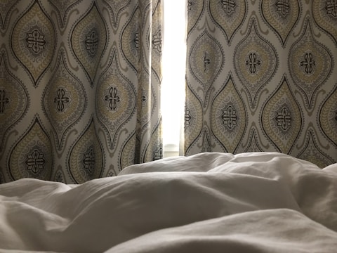 A softly lit bedroom with patterned curtains, featuring an intricate design of overlapping ovals and floral elements. The light from the window is partially obscured by the curtains, casting a gentle glow onto the rumpled white bedding in the foreground.