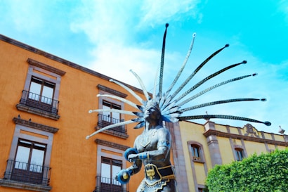 A statue of an Indigenous figure with elaborate headdress stands prominently against the backdrop of a colorful building. The building has multiple windows with balconies and features warm orange and yellow tones. The bright blue sky enhances the vibrancy of the scene.