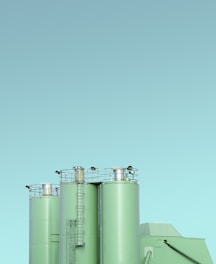 Three industrial silos with a minimalist design, featuring a light green color against a clear blue sky. The silos have visible ladders and metal railings on top, with clean lines and a modern appearance.