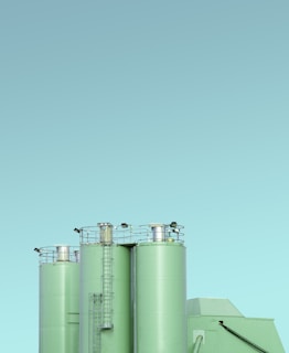 Three industrial silos with a minimalist design, featuring a light green color against a clear blue sky. The silos have visible ladders and metal railings on top, with clean lines and a modern appearance.