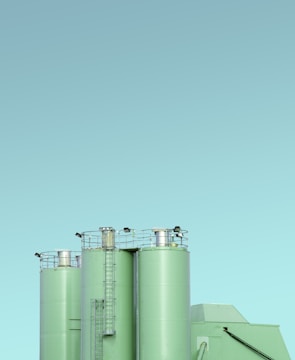Three industrial silos with a minimalist design, featuring a light green color against a clear blue sky. The silos have visible ladders and metal railings on top, with clean lines and a modern appearance.