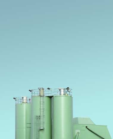 Three industrial silos with a minimalist design, featuring a light green color against a clear blue sky. The silos have visible ladders and metal railings on top, with clean lines and a modern appearance.