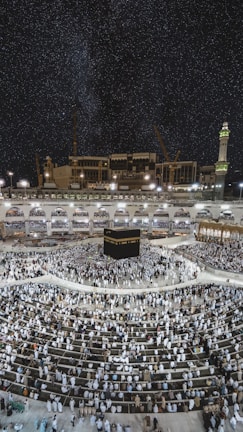 A large crowd of people gathered around the Kaaba, a cuboidal structure located at the center of the Grand Mosque in Mecca, Saudi Arabia. The image captures the scene at night, under a clear, starry sky. The mosque is surrounded by illuminated structures and features a prominent minaret on the right.