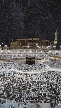 A large crowd of people gathered around the Kaaba, a cuboidal structure located at the center of the Grand Mosque in Mecca, Saudi Arabia. The image captures the scene at night, under a clear, starry sky. The mosque is surrounded by illuminated structures and features a prominent minaret on the right.