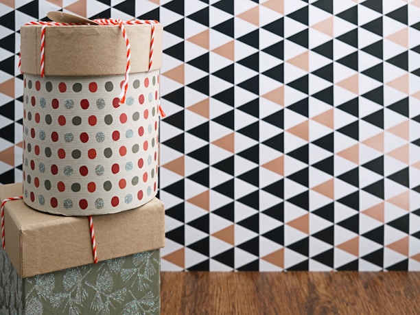 A patterned cylindrical box with a lid, adorned with red and silver polka dots, is stacked on a square box with a kraft paper appearance. A red and white string is placed on top. The background features a geometric black and beige triangle pattern on the wall, with a wooden floor beneath.