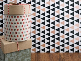 A patterned cylindrical box with a lid, adorned with red and silver polka dots, is stacked on a square box with a kraft paper appearance. A red and white string is placed on top. The background features a geometric black and beige triangle pattern on the wall, with a wooden floor beneath.