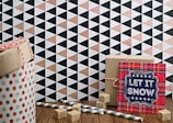 A gift-wrapped cylindrical container with polka dots and a red and white twisted cord sits on a wooden surface. Nearby, there is a rectangular box tied with red string and a card with a plaid pattern reading 'Let It Snow' in bold letters. Sugar cubes and striped paper straws are scattered around, and the background features a geometric pattern of triangles in black, white, and peach tones.
