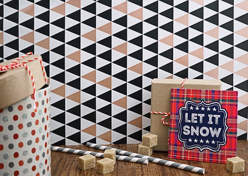 A gift-wrapped cylindrical container with polka dots and a red and white twisted cord sits on a wooden surface. Nearby, there is a rectangular box tied with red string and a card with a plaid pattern reading 'Let It Snow' in bold letters. Sugar cubes and striped paper straws are scattered around, and the background features a geometric pattern of triangles in black, white, and peach tones.