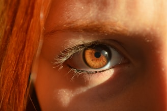 A close-up of an eye with striking amber colors, surrounded by long eyelashes and framed by reddish-brown hair. The lighting emphasizes the iris, highlighting its intricate patterns.