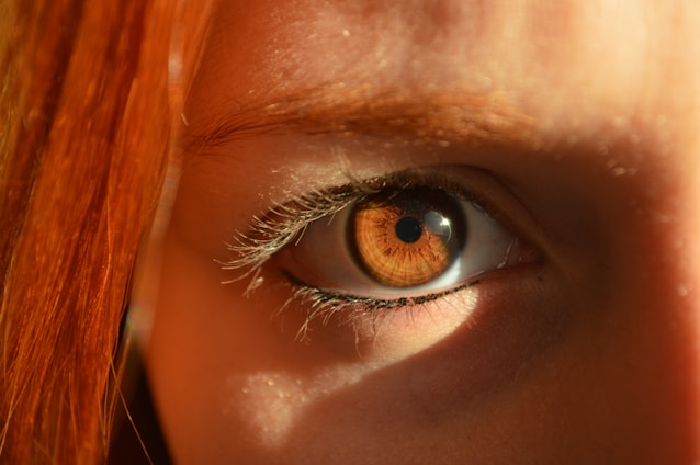 A close-up of an eye with striking amber colors, surrounded by long eyelashes and framed by reddish-brown hair. The lighting emphasizes the iris, highlighting its intricate patterns.
