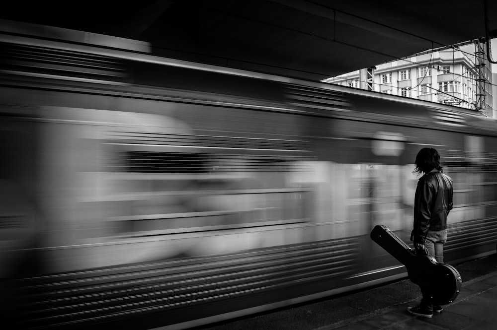 기차 앞에 서 있는 기타 케이스를 들고 있는 남자의 회색조 사진
