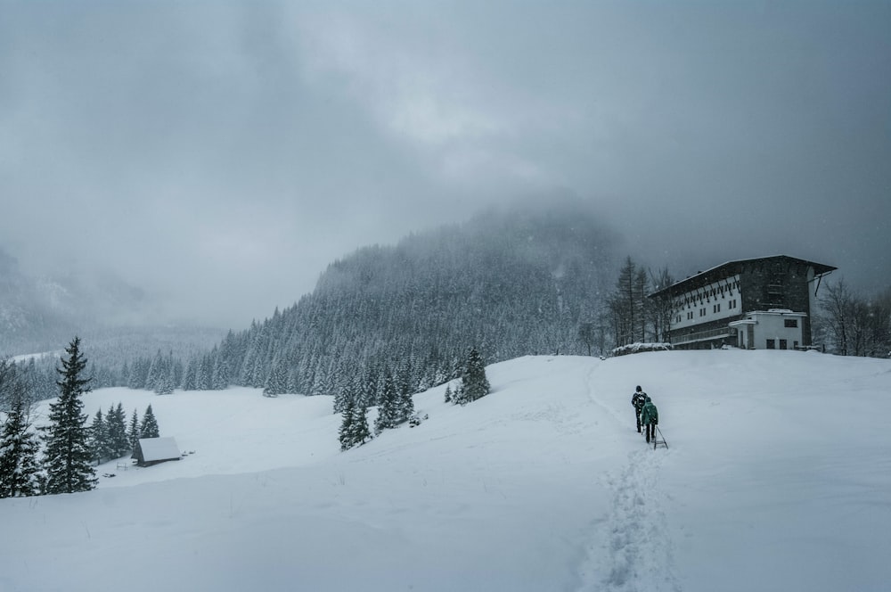 two persons walking on snow near concrete house under nimbus clouds