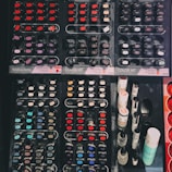A display of various nail polish bottles arranged in an organized manner. The layout includes multiple rows of small, circular and oval nail polish containers featuring a wide spectrum of colors such as reds, blues, purples, and blacks. On the right side of the display, taller bottles of nail products are visible, possibly including top coats or related items.