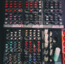 A display of various nail polish bottles arranged in an organized manner. The layout includes multiple rows of small, circular and oval nail polish containers featuring a wide spectrum of colors such as reds, blues, purples, and blacks. On the right side of the display, taller bottles of nail products are visible, possibly including top coats or related items.
