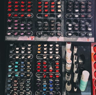 A display of various nail polish bottles arranged in an organized manner. The layout includes multiple rows of small, circular and oval nail polish containers featuring a wide spectrum of colors such as reds, blues, purples, and blacks. On the right side of the display, taller bottles of nail products are visible, possibly including top coats or related items.