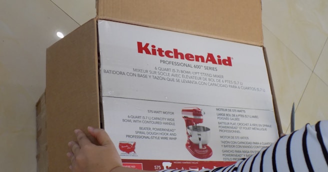 A person is opening a cardboard box that contains a KitchenAid Professional 600 series stand mixer. The box is labeled with product details in multiple languages and shows an image of a red stand mixer. The person’s hand is holding the box, and they are wearing a striped shirt.