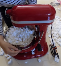 A red stand mixer is filled with white packing peanuts. A person wearing a striped shirt is adjusting the mixer. Packing peanuts are scattered around the floor.