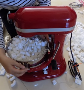 A red stand mixer is filled with white packing peanuts. A person wearing a striped shirt is adjusting the mixer. Packing peanuts are scattered around the floor.