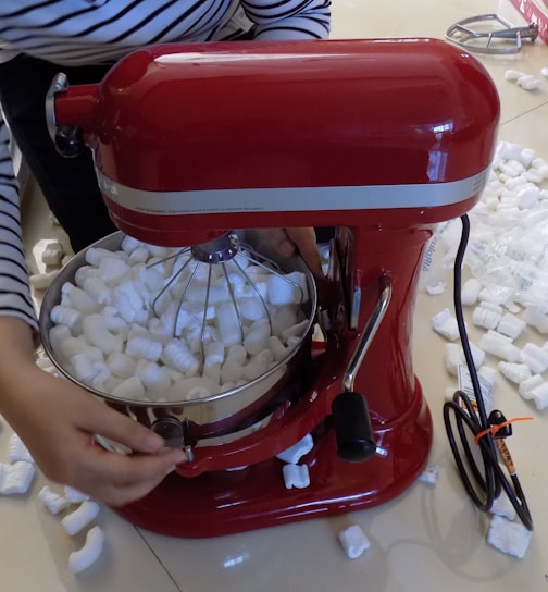 A red stand mixer is filled with white packing peanuts. A person wearing a striped shirt is adjusting the mixer. Packing peanuts are scattered around the floor.