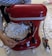 A red stand mixer is filled with white packing peanuts. A person wearing a striped shirt is adjusting the mixer. Packing peanuts are scattered around the floor.