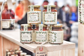 Several jars of curry paste and chutney are displayed on a wooden shelf. The labels feature decorative borders with text such as 'East Yorkshire Piccalilli', 'Ruby Curry Paste', and 'Mango, Methi & Fruit Chutney'. The jars are neatly arranged, and more bottles can be seen in the background amidst a blurred crowd.