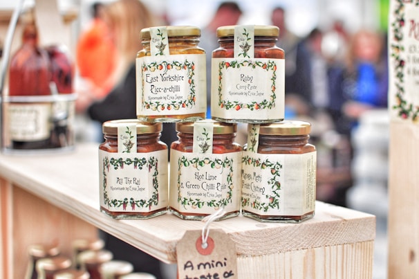 Several jars of curry paste and chutney are displayed on a wooden shelf. The labels feature decorative borders with text such as 'East Yorkshire Piccalilli', 'Ruby Curry Paste', and 'Mango, Methi & Fruit Chutney'. The jars are neatly arranged, and more bottles can be seen in the background amidst a blurred crowd.