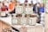 Several jars of curry paste and chutney are displayed on a wooden shelf. The labels feature decorative borders with text such as 'East Yorkshire Piccalilli', 'Ruby Curry Paste', and 'Mango, Methi & Fruit Chutney'. The jars are neatly arranged, and more bottles can be seen in the background amidst a blurred crowd.